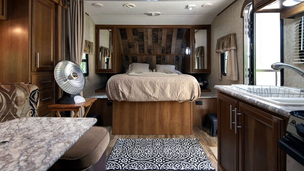 A compact RV bedroom with a bed, a small table, a fan, and a kitchenette.