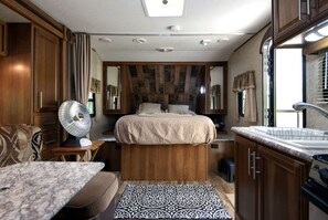 Een compacte slaapkamer in een camper, met een bed, een klein tafeltje, een ventilator en een kitchenette.