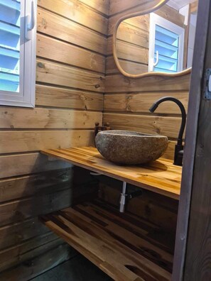 A wooden cabin interior with a stone sink, wooden shelves, and a window with blinds. - Bungalow (Sainte-Anne)