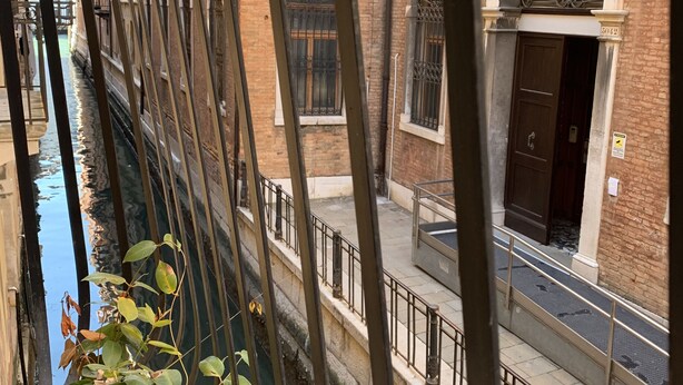 A narrow canal with a metal railing, a potted plant, and a building with a large door and windows. - Three bedroom apartment in Venice San Marco (Venezia)
