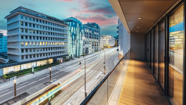 A modern building with a balcony overlooking a city street and train tracks.