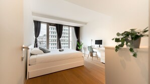 A modern bedroom with a bed, a desk, and a view of buildings through large windows.