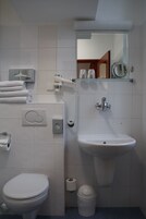 Casa de banho com sanita, lavatório e espelho. Toalhas e um contentor de lixo estão visíveis.