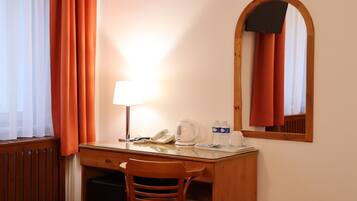 Quarto de hotel com uma escrivaninha de madeira, uma cadeira, abajur, telefone, chaleira e espelho.