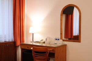 Quarto de hotel com uma escrivaninha de madeira, uma cadeira, abajur, telefone, chaleira e espelho.