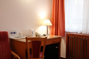 Quarto de hotel com escrivaninha de madeira, cadeira, abajur, telefone, chaleira e radiador.