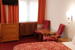 Quarto de hotel com cama, cómoda de madeira, mesa, duas cadeiras vermelhas, radiador e TV fixada na parede.