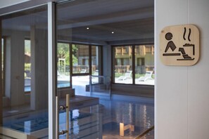A modern hotel lobby with a glass entrance, a wooden sign indicating a sauna area, and a view of the outdoor pool and lounge chairs.