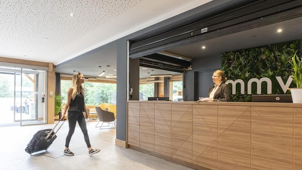 Un espace d’accueil comprenant un comptoir de réception en bois, un(e) réceptionniste et une personne tenant une valise.