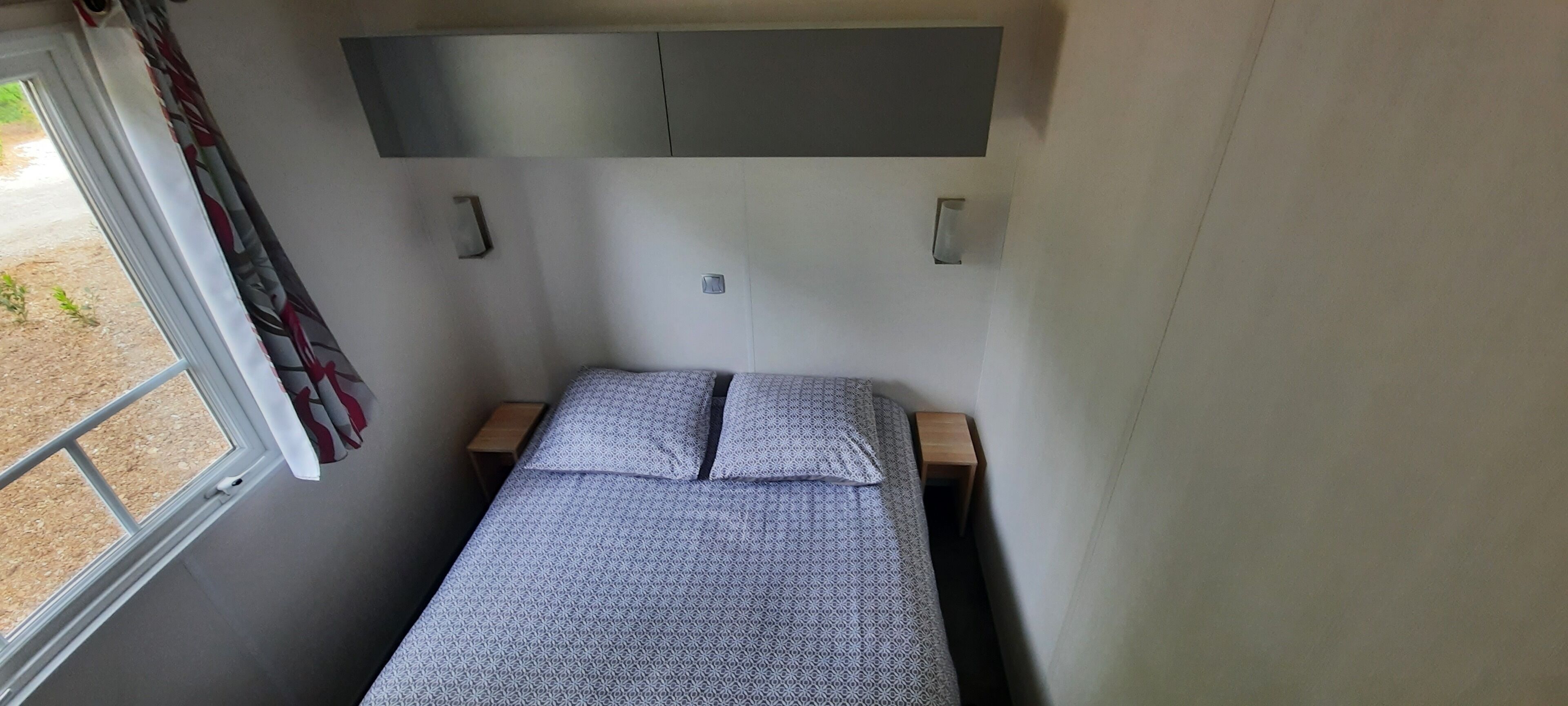 A small, compact bedroom with a single bed, a window with curtains, and a built-in shelf above the bed.