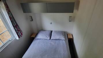 A small, compact bedroom with a single bed, a window with curtains, and a built-in shelf above the bed.