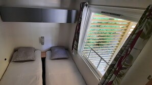 A small, compact room with two single beds, a window with blinds, and a wall-mounted air conditioning unit. - Mobil home 6 pers camping 3* sur belle parcelle au calme avec piscine (Saint-Chéron)