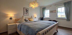 Un dormitorio con cama, mesitas de noche, lámpara de pared, cuadro enmarco y ventana con cortinas.