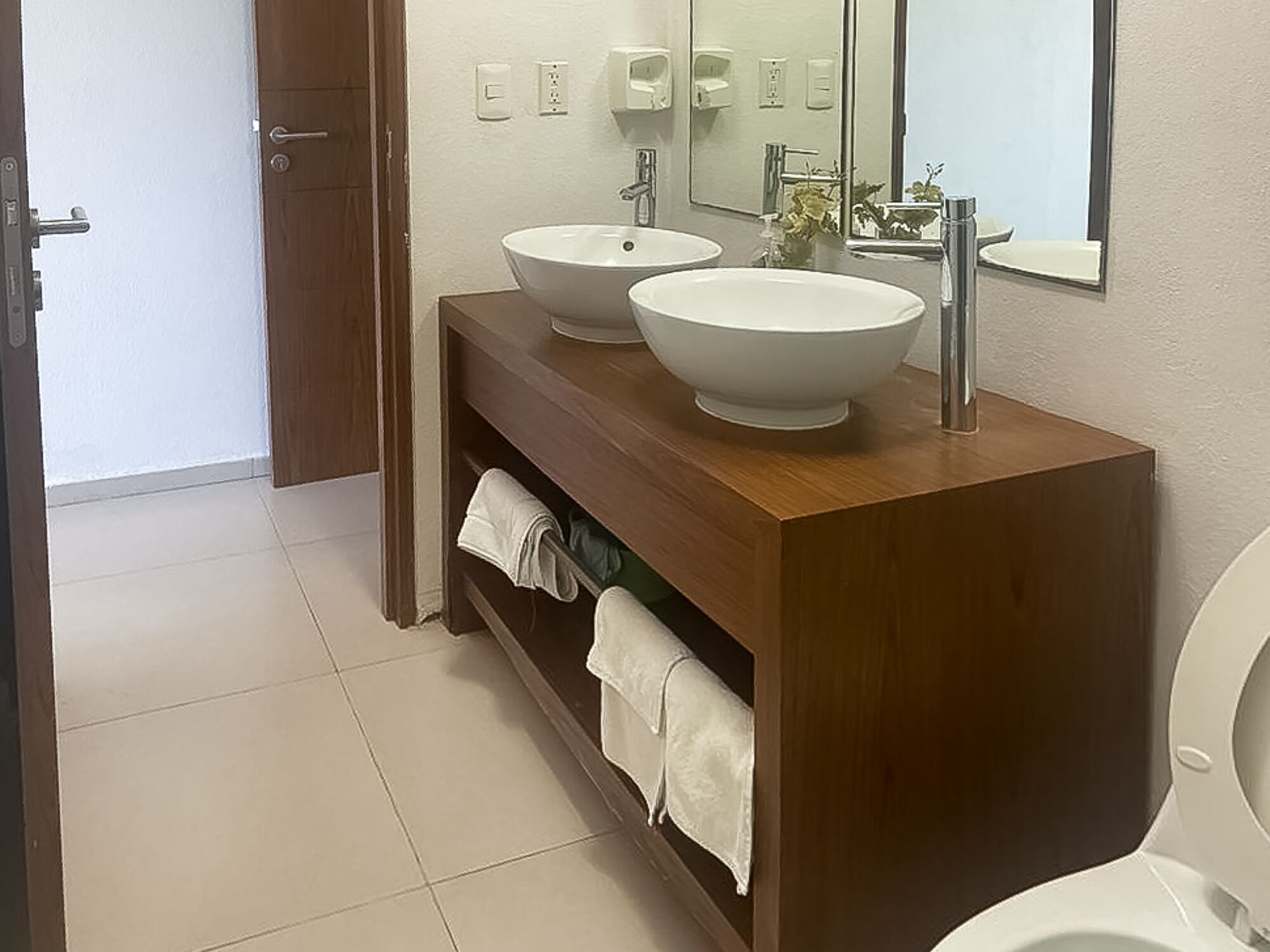 Een badkamer met twee witte wasbakken, een houten wastafelmeubel, een spiegel en een toilet.