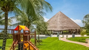 A playground with a slide and a thatched-roof structure in a grassy area with palm trees. - AMARA Beachfront Apartments by The Sea (Villa de Cos)