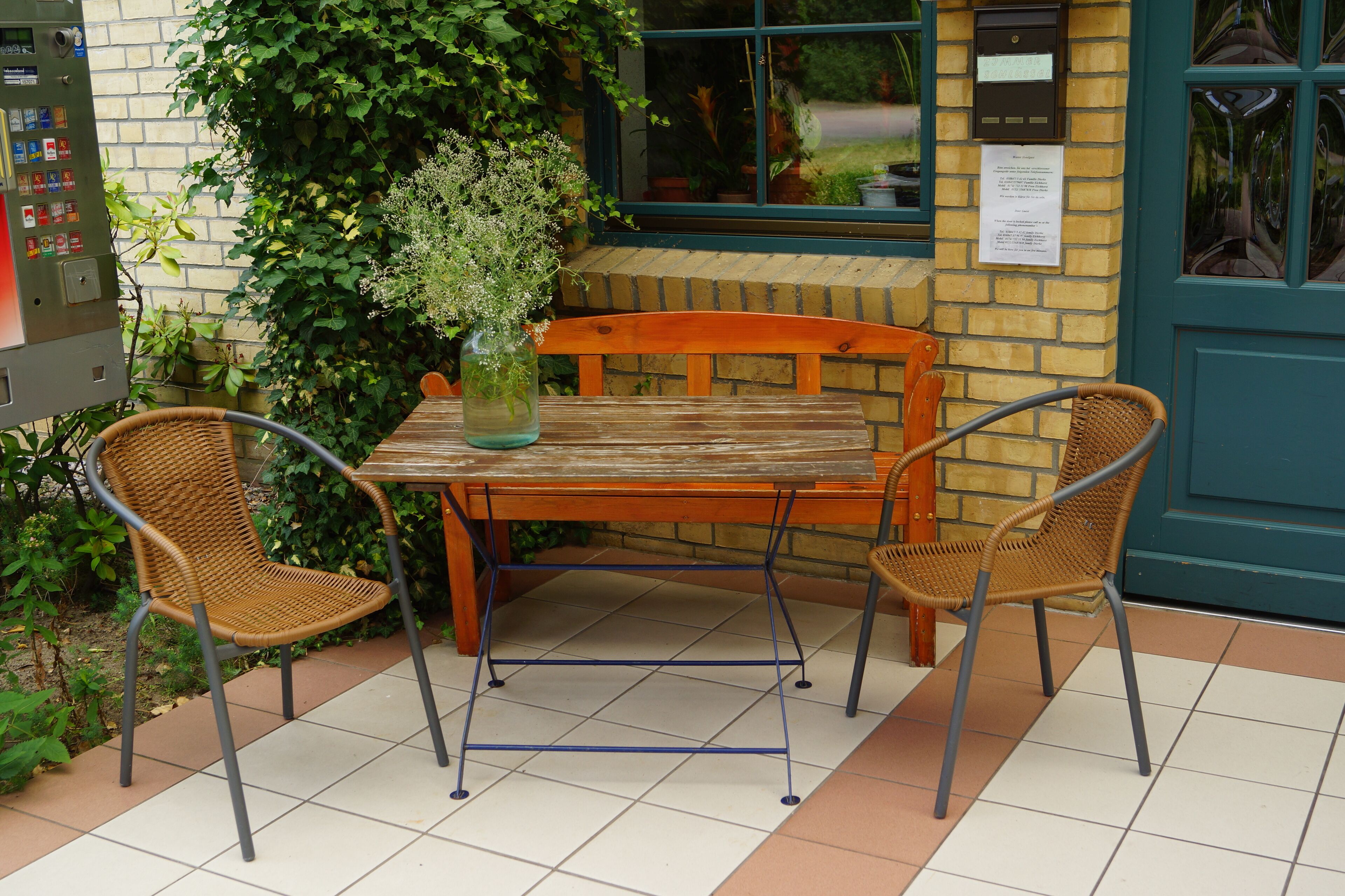 An outdoor seating area with a wooden table and wicker chairs, surrounded by greenery and a brick wall.