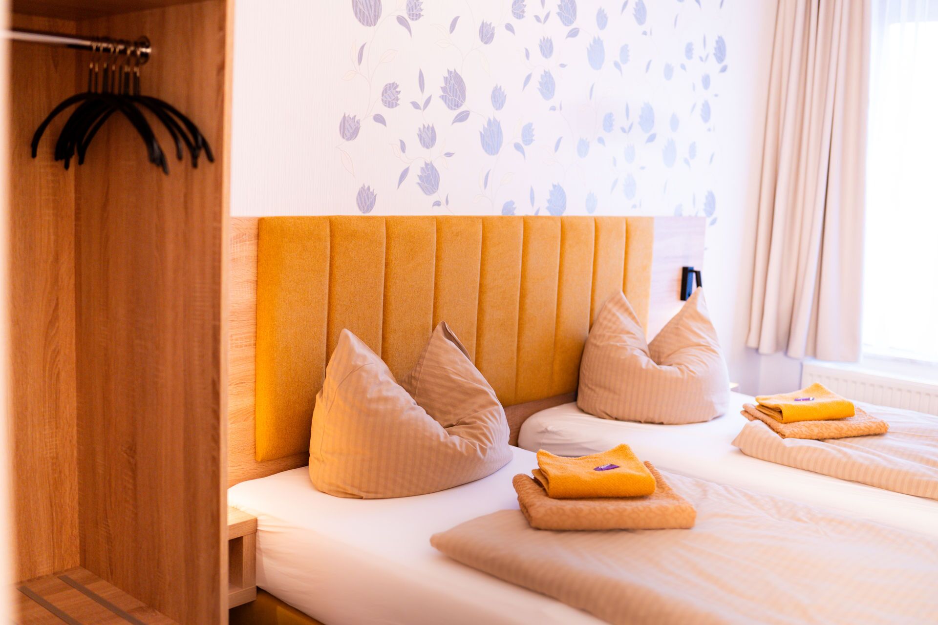 Quarto de hotel com guarda-roupa de madeira, cabeceira com padrão floral e cama com almofadas bege e toalha dobrada.
