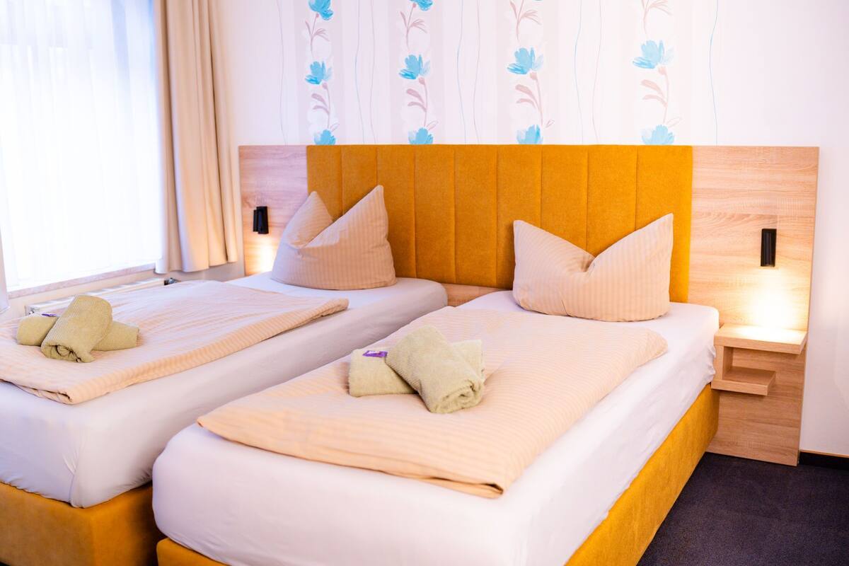 Une chambre d’hôtel avec deux lits, chacun doté d’une tête de lit, d’oreillers et de serviettes. Il y a une table de chevet avec une lampe de chaque côté des lits.