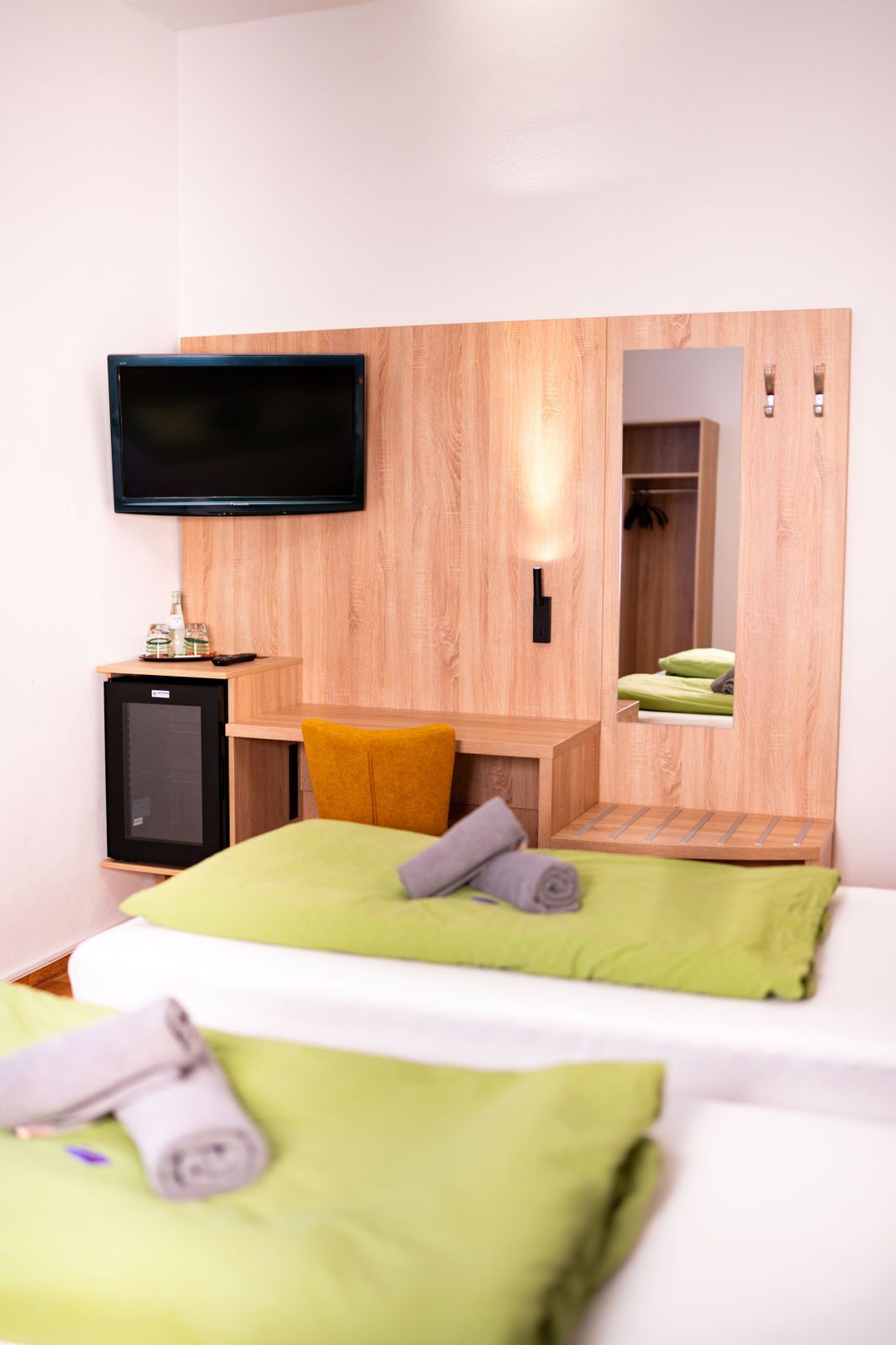 Quarto de hotel com cama, televisão fixada na parede, pequena escrivaninha e guarda-roupa.