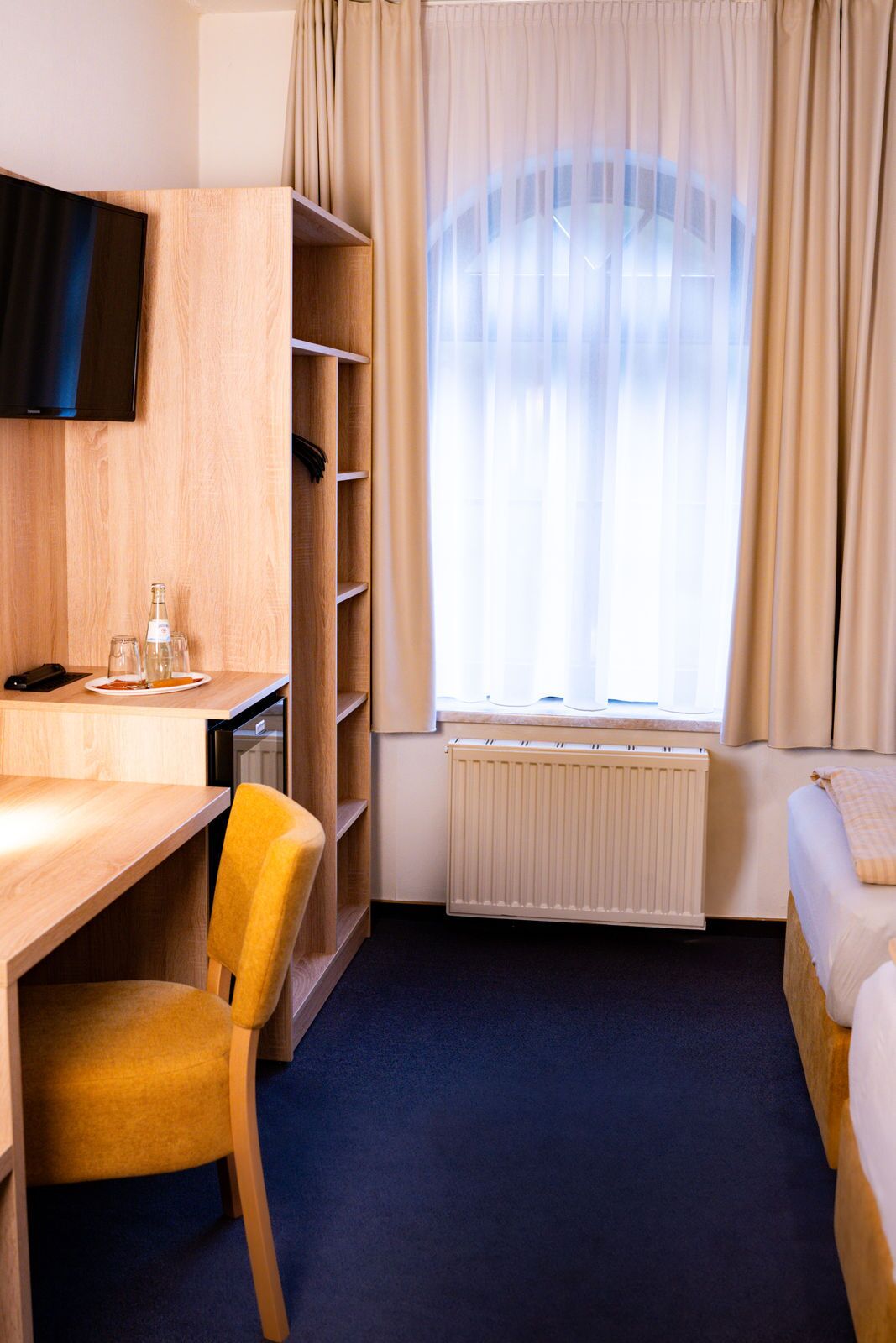 Hotellihuone, jossa on puinen työpöytä, tuoli, sänky, seinälle asennettu televisio, ikkuna verhoilla ja patteri.
