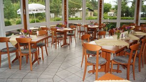 Een restaurant met meerdere tafels en stoelen, grote ramen en uitzicht op groen.