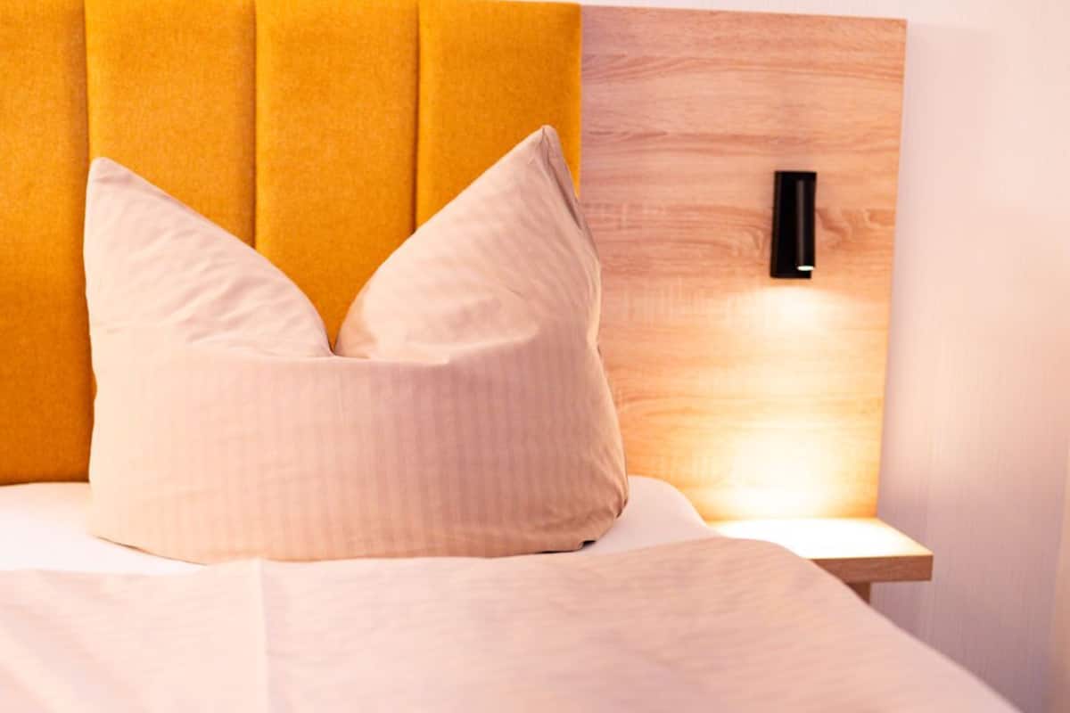 Un lit bien fait, avec une tête de lit jaune et deux oreillers, une table de chevet en bois avec une lampe, et un luminaire fixé au mur.