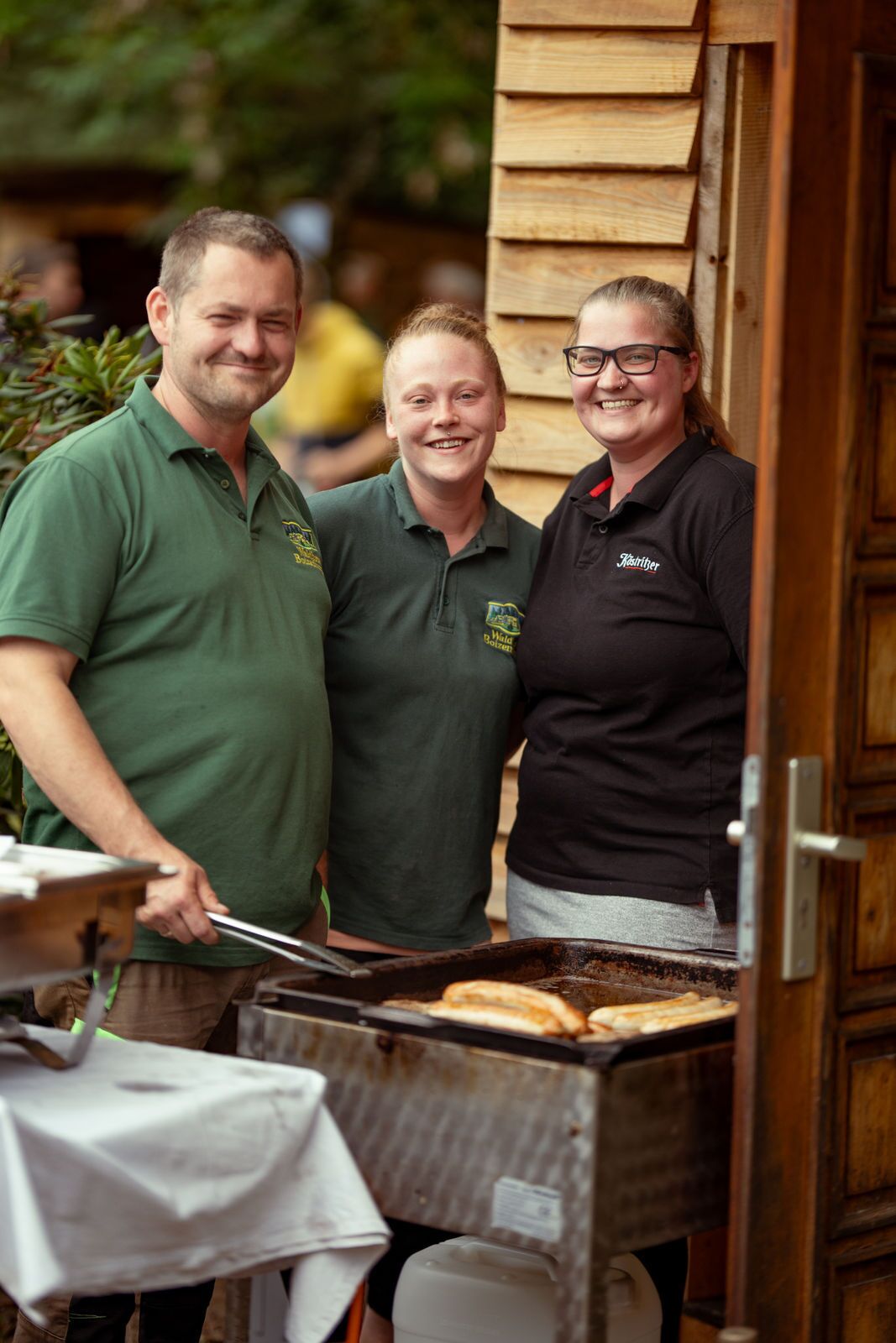 Tres personas de pie frente a una parrilla con comida cocinándose.
