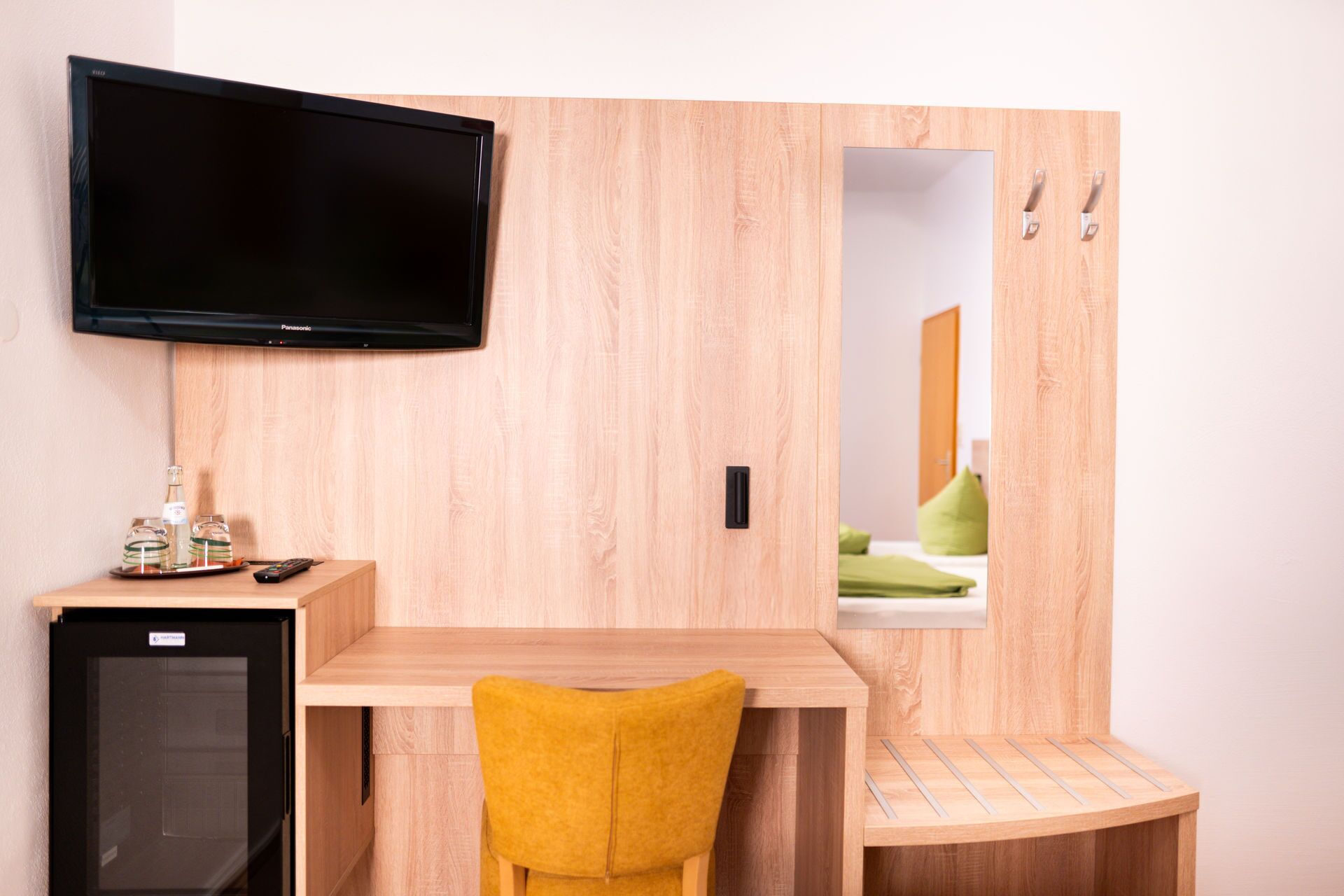A compact hotel room with a wooden desk, a small refrigerator, a flat-screen TV, a mirror, and a yellow chair.
