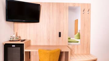 A compact hotel room with a wooden desk, a small refrigerator, a flat-screen TV, a mirror, and a yellow chair.