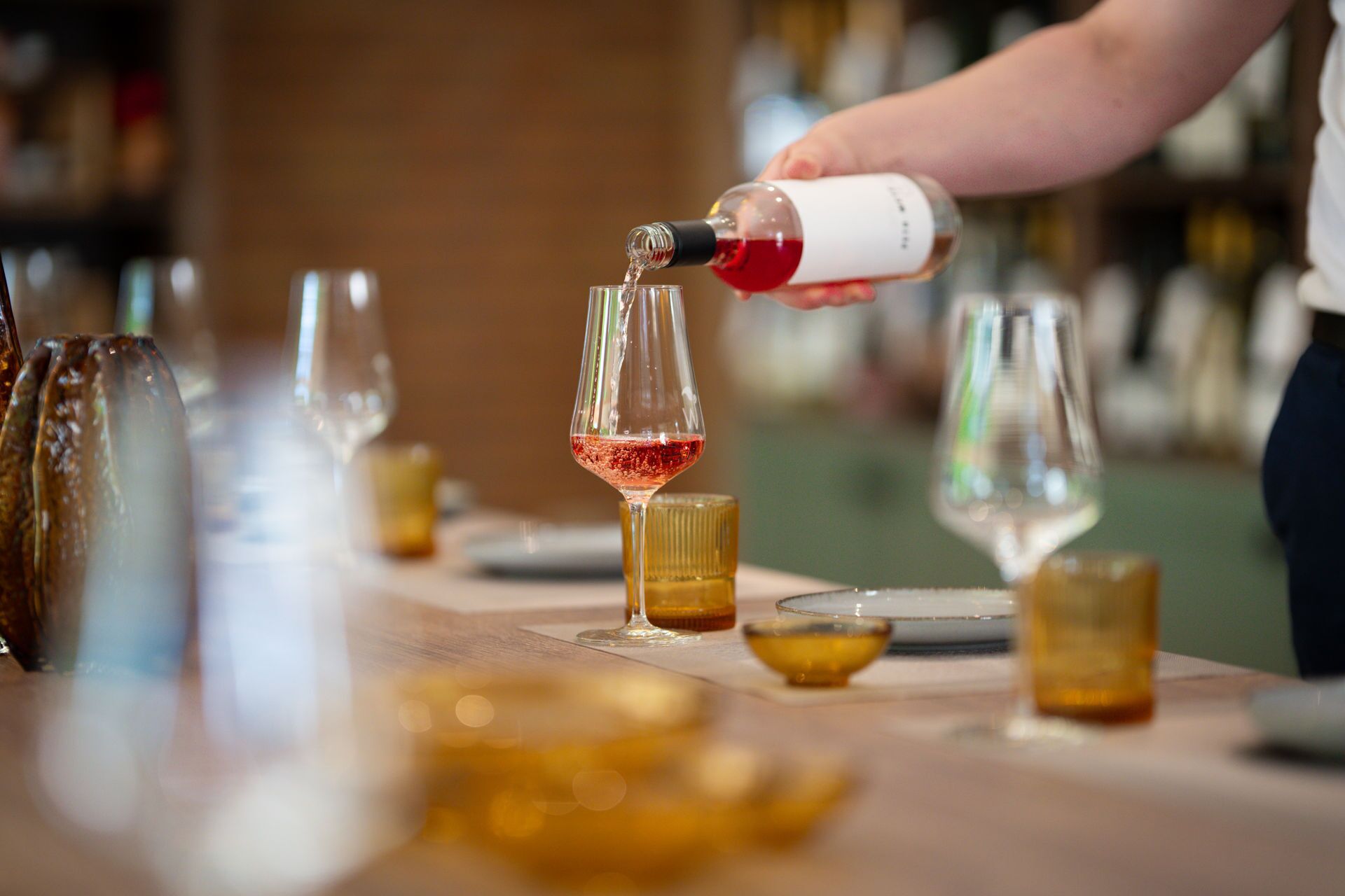A person pouring red wine into a glass on a bar counter with multiple glasses and bottles in the background.