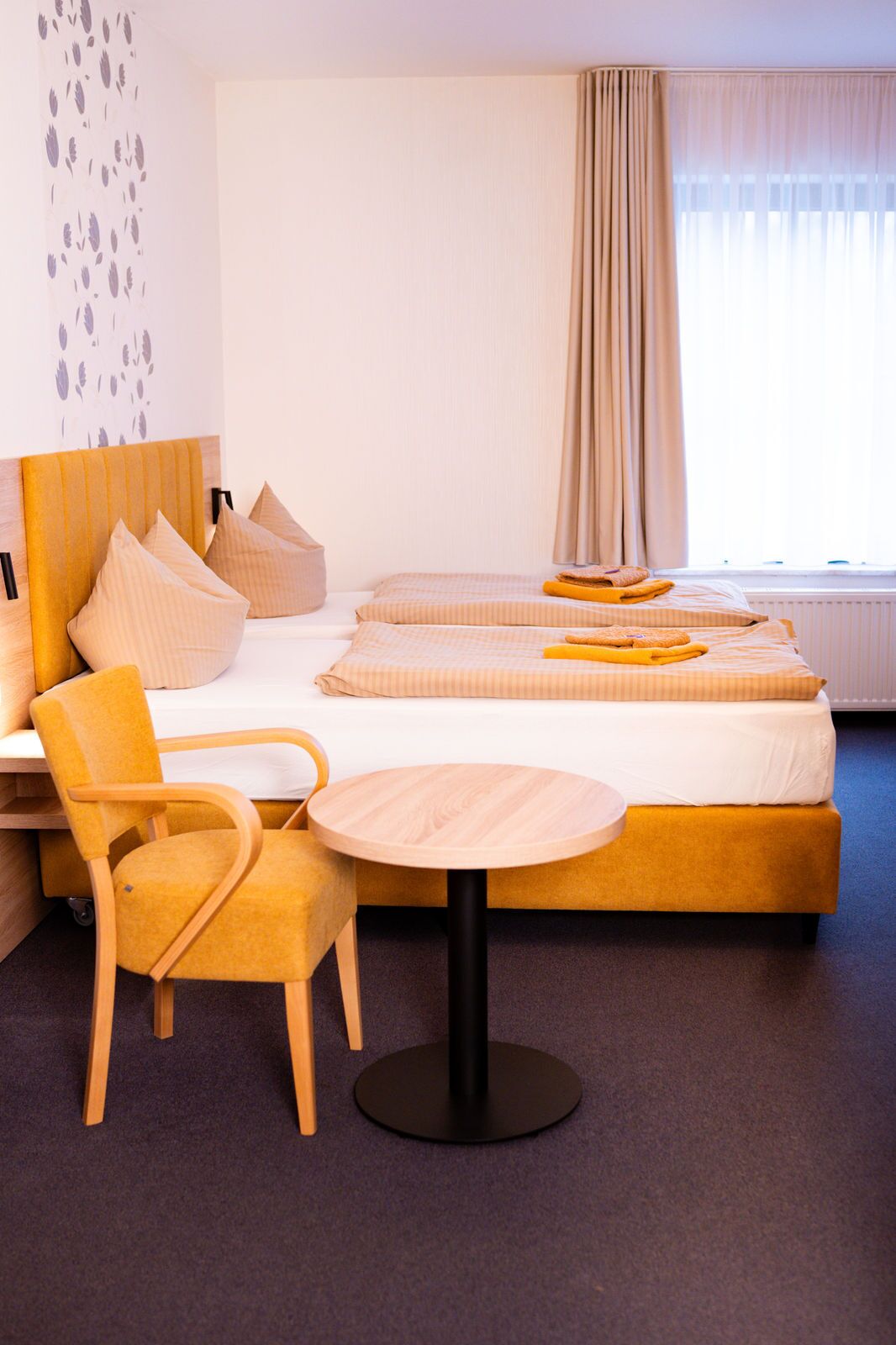 A hotel room with a bed, two pillows, a chair, a small round table, and a window with curtains.