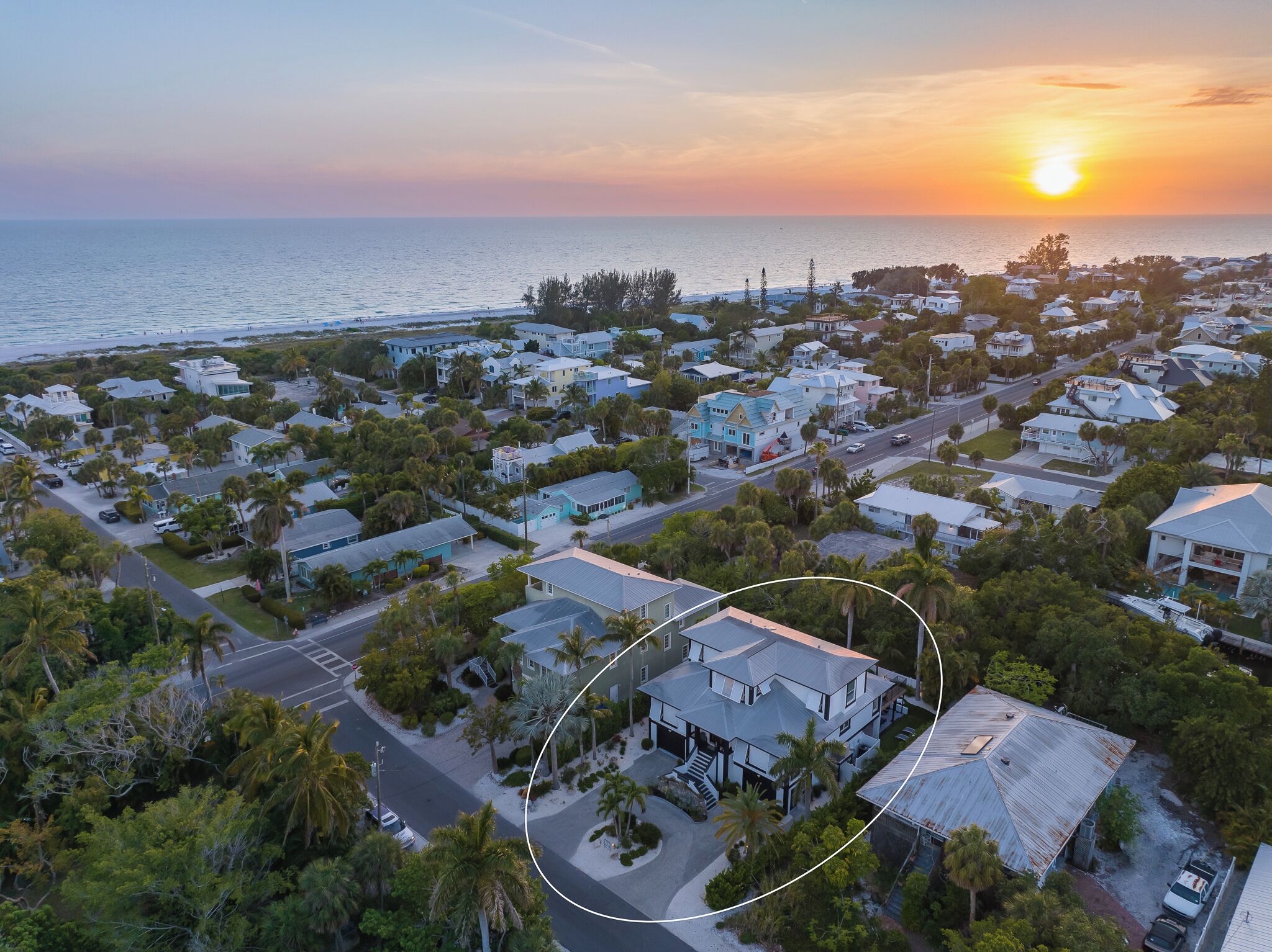 Vue aérienne d’un quartier résidentiel situé sur la côte, avec des maisons, des palmiers et un coucher de soleil sur l’océan.
