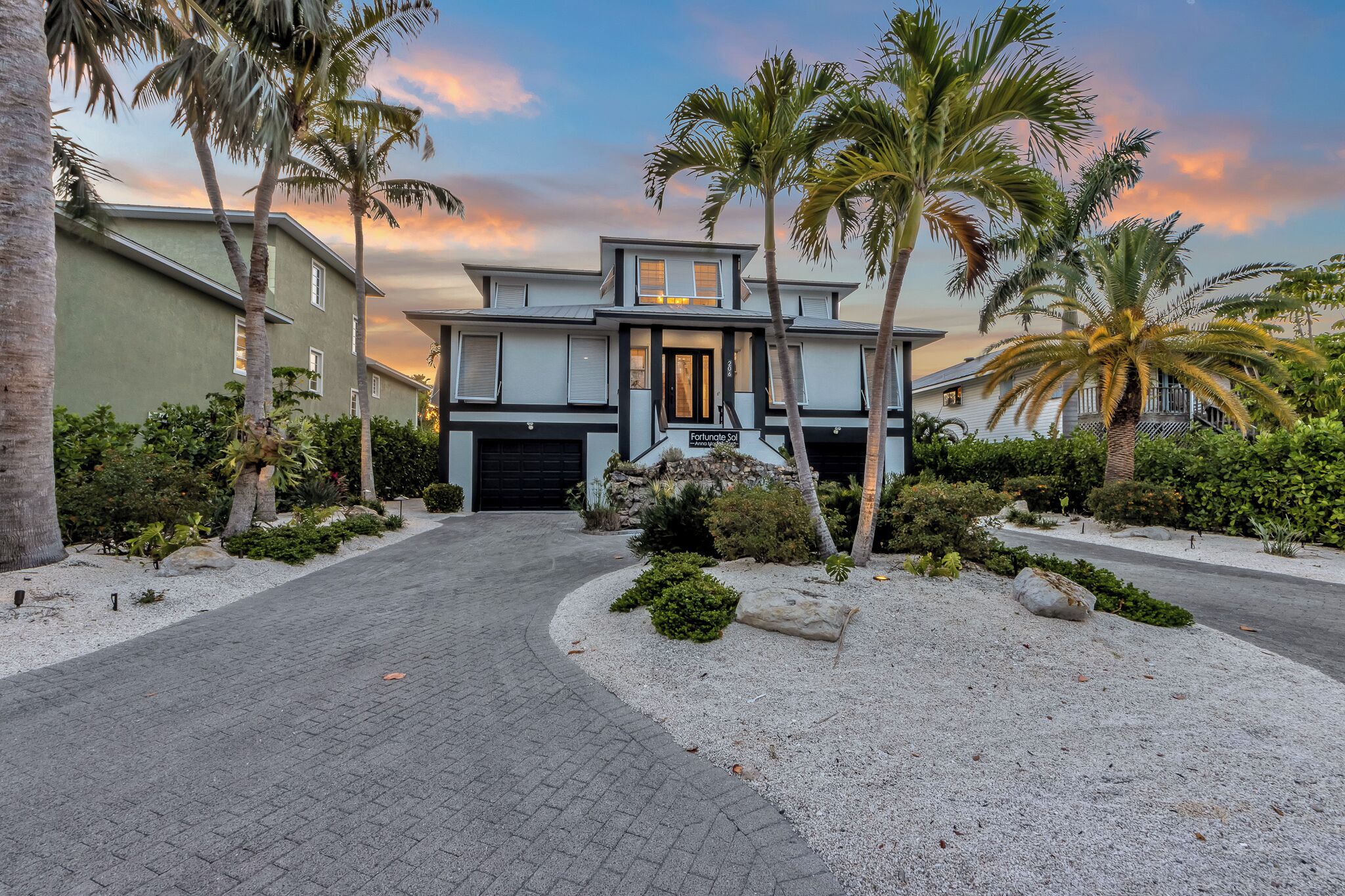 A modern two-story house with a driveway leading to a garage, surrounded by palm trees and well-maintained landscaping.