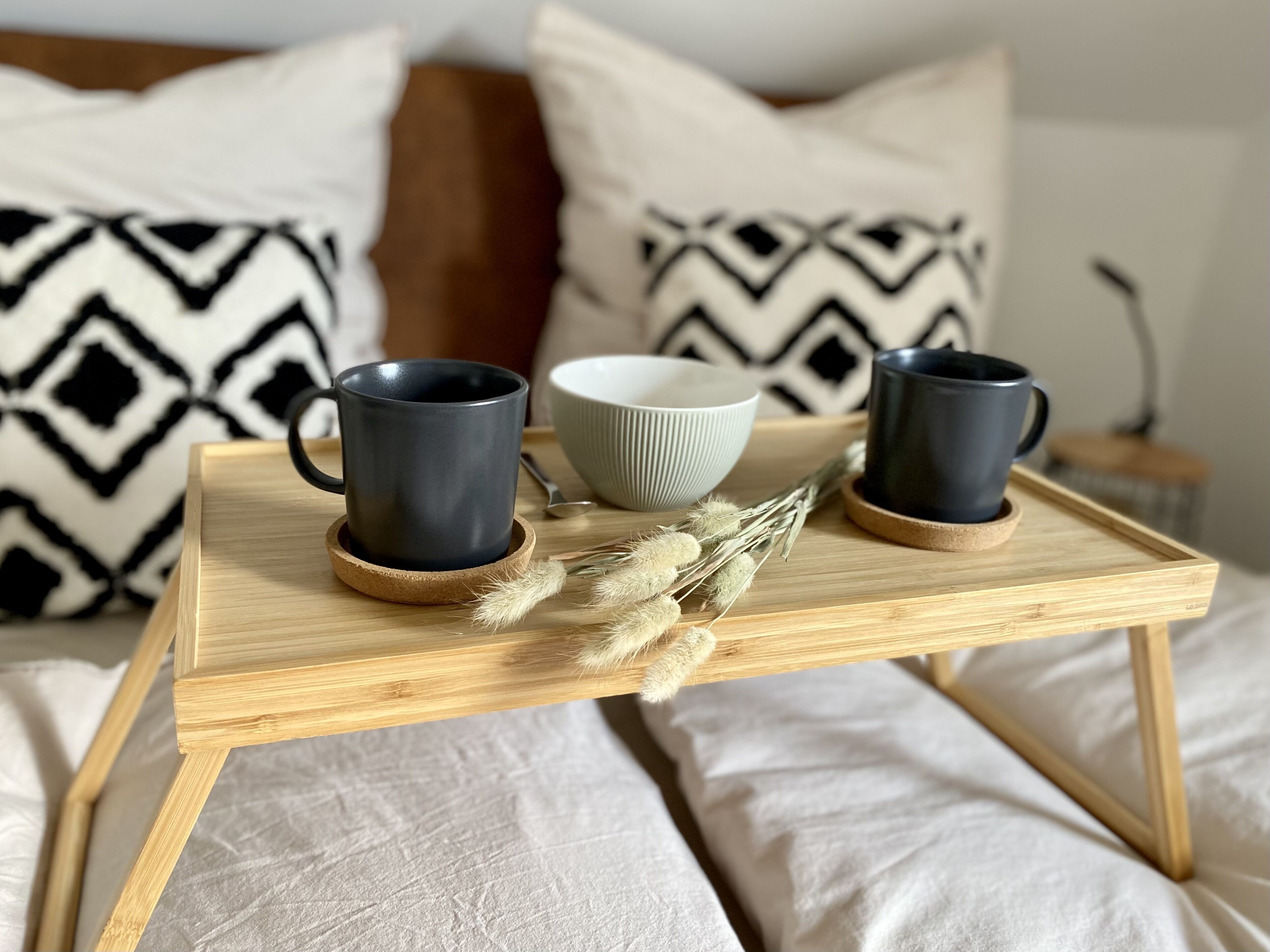 Un plateau en bois sur lequel reposent deux tasses foncées et un bol blanc, le tout posé sur un lit agrémenté d’oreillers à motifs.