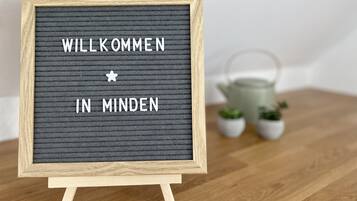 A framed welcome sign with the text "WILLKOMMEN IN MINDEN" on a textured background, placed on a wooden surface with small potted plants in the background.