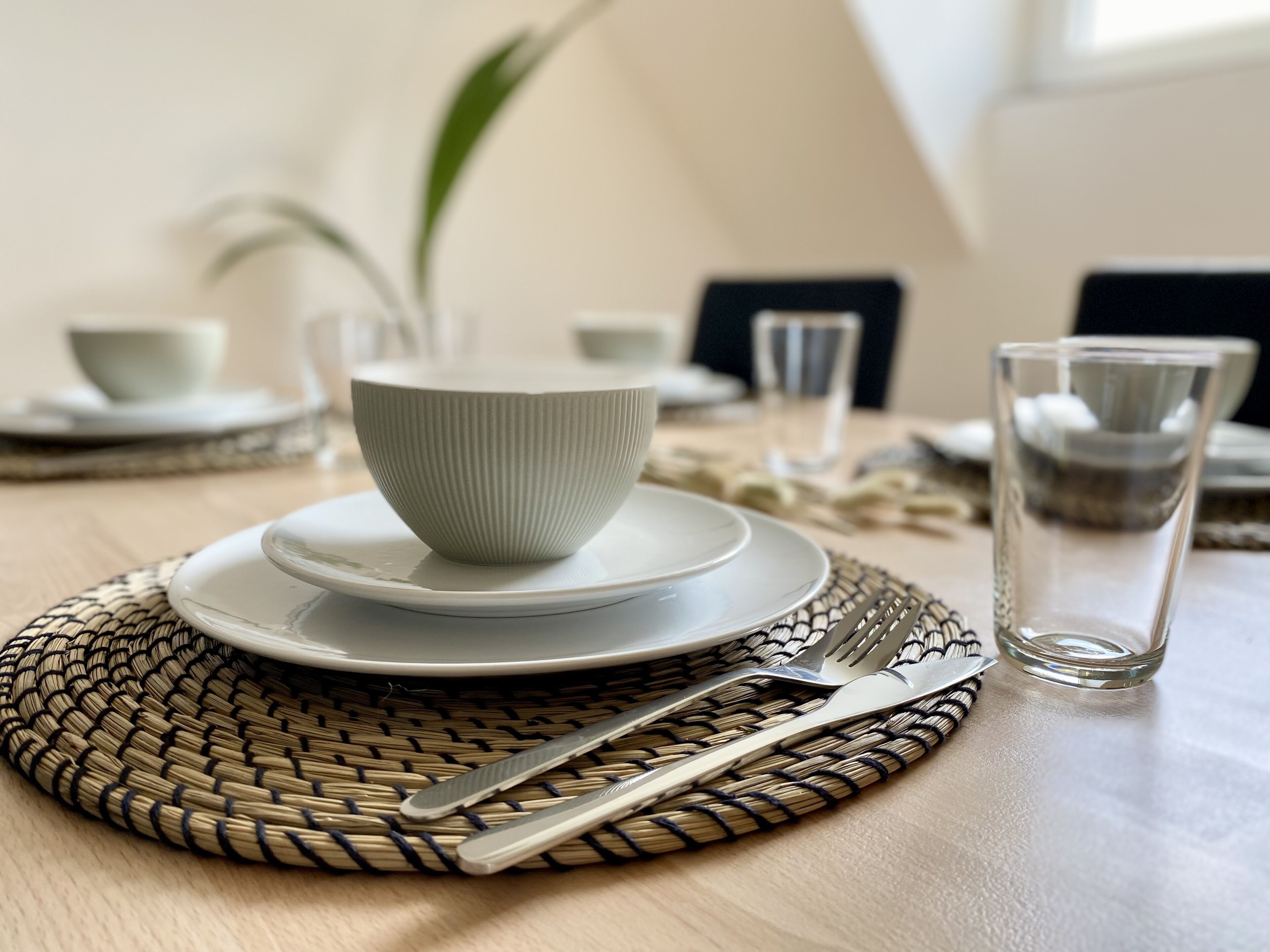 Une table à manger dressée avec des assiettes blanches, un bol, des couverts et un verre.
