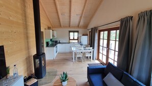 A cozy cabin interior with a wooden floor, a dining area with a table and chairs, a kitchen with white cabinets, and a large window with curtains. - Ecological new-build wooden block vacation home - free AlbCard from 2025  (Hayingen)