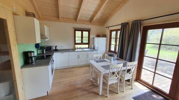 Eine kompakte Küche mit weißen Schränken, einer Kaffeemaschine, einer Mikrowelle und einem Spülbecken. Ein Esstisch mit Stühlen und ein Fenster mit Vorhängen sind ebenfalls vorhanden.