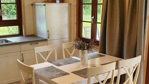 A dining area with a white table and chairs, a vase with flowers, and a window with curtains. - Ecological new-build wooden block vacation home - free AlbCard from 2025  (Hayingen)