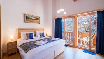 A bedroom with a large bed, a wooden headboard, a nightstand with a lamp, a painting on the wall, and a balcony with a view of trees.