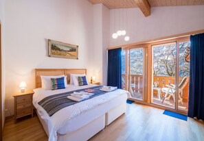 A bedroom with a large bed, a wooden headboard, a nightstand with a lamp, a painting on the wall, and a balcony with a view of trees. - Racers Retreat 3 (Wengen)