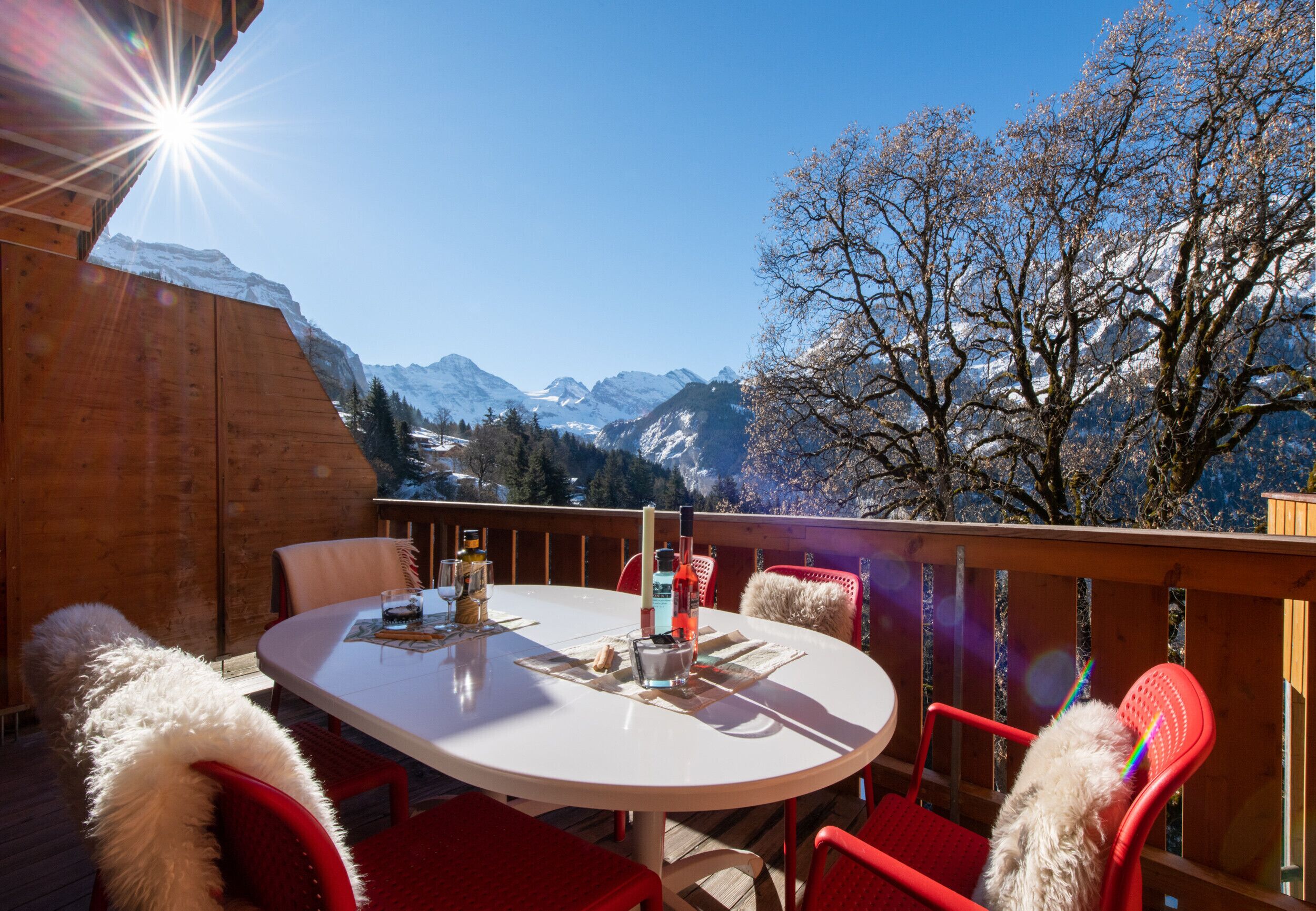 A round table with chairs on an outdoor deck, overlooking snow-capped mountains and a clear blue sky.