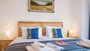 A neatly made double bed with a striped bedspread, two pillows with a colorful pattern, and a bottle of wine on a bedside table.