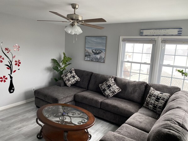 A modern living room with a sectional sofa, a glass coffee table, a ceiling fan, and a large window with blinds.