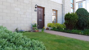 Un edificio con facciata in mattoni, una porta in legno e un piccolo giardino con piante e fiori in vaso.