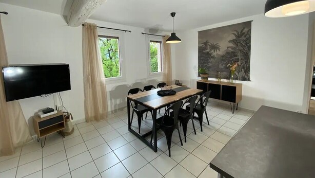 A modern living room with a dining area, large windows, a flat-screen TV, and a painting on the wall.