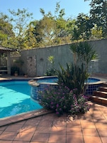 Una piscina con borde de azulejos, rodeada por un muro de piedra y un pequeño jardín con flores púrpuras.