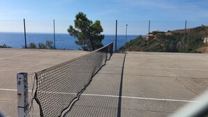 An empty tennis court with a chain-link fence, a metal post, and a view of the sea and hills in the background.