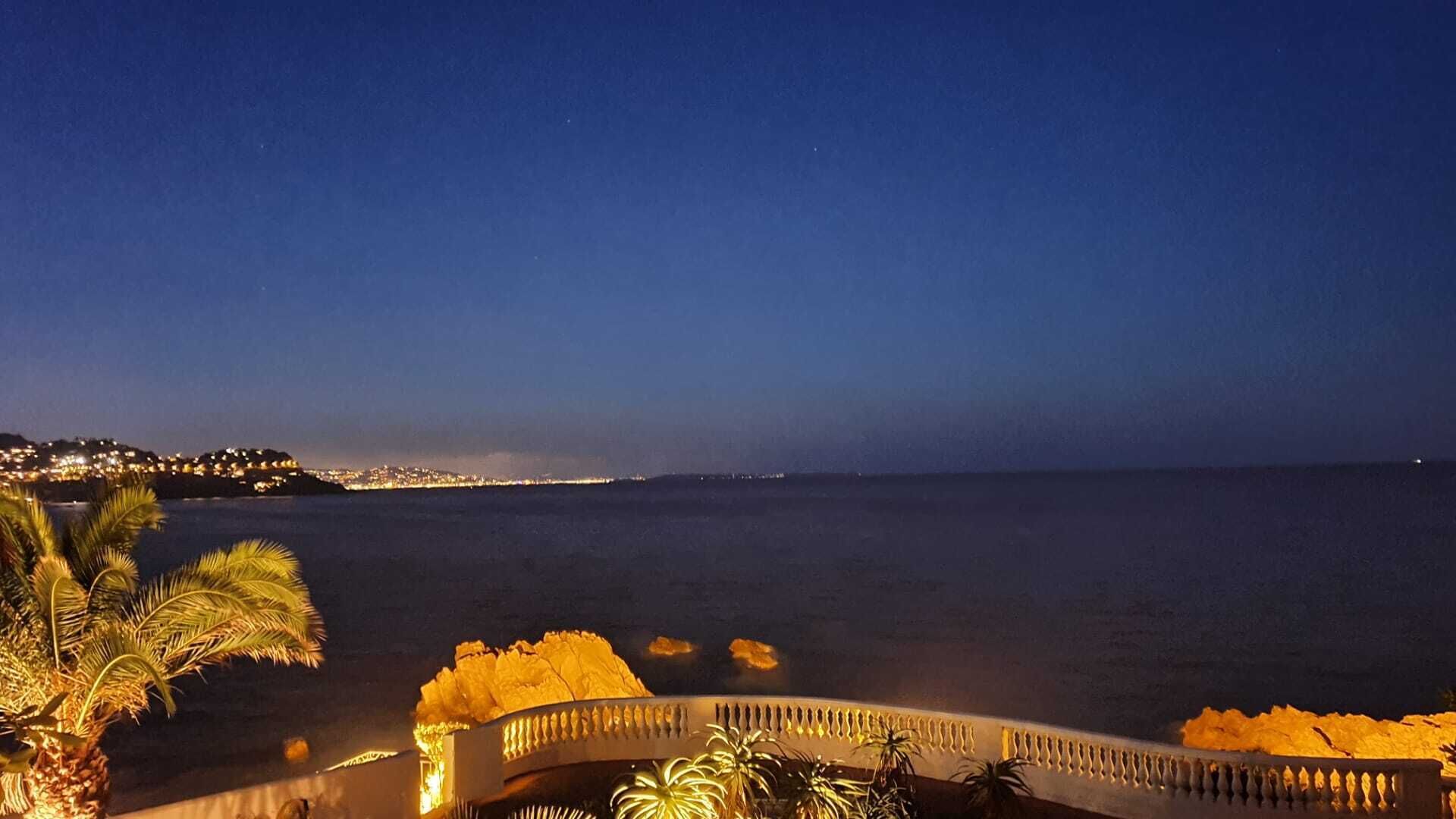 Vista costera con una playa iluminada y un paisaje urbano distante.