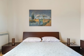 Quarto com cama, duas almofadas, mesa de cabeceira com abajur e quadro na parede.